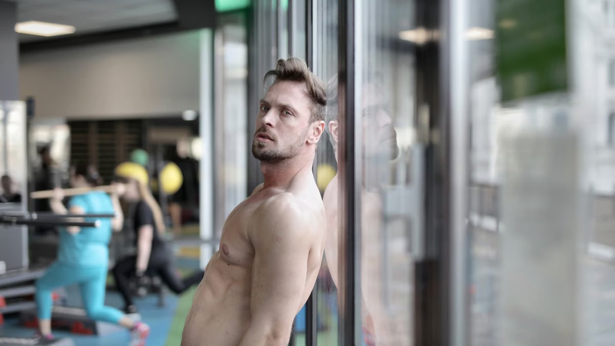 Shirtless man reflects at gym window, showcasing muscular physique and determination.